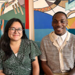photo of nayeli wearing glasses and green polka dot top sitting on left and ryan wearing collared cream color layered shirts on right, in front of mural in LGF office