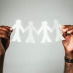 person with brown skin holds a paper chain of 4 human shapes between their hands