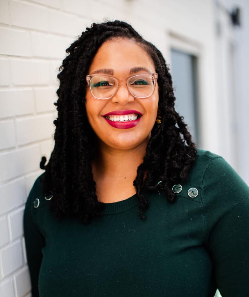 headshot of marion johnson - wearing a dark green shirt and glasses with white brick background