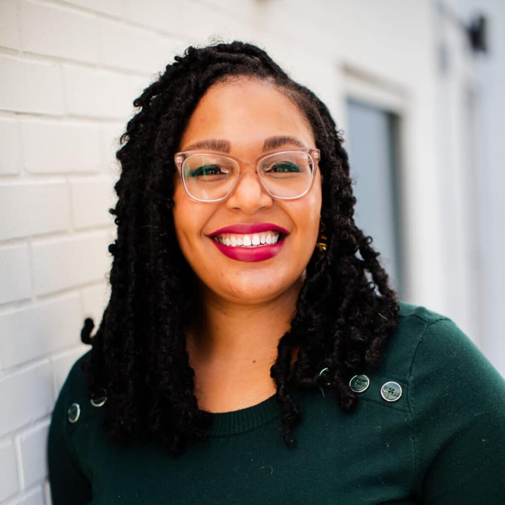 headshot of marion johnson - wearing a dark green shirt and glasses with white brick background