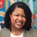 ebony west headshot - ebony is wearing a white shirt with grey blazer in front of a multicolor mural backdrop