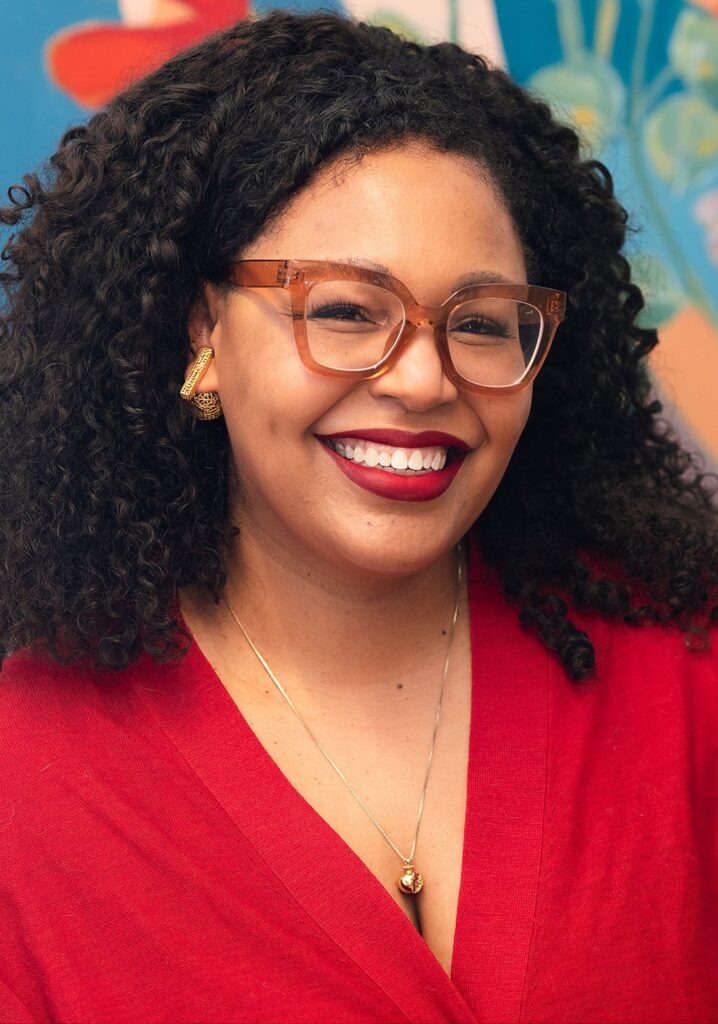 Marion Johnson headshot - She's wearing red glasses and a red shirt against a mural backdrop